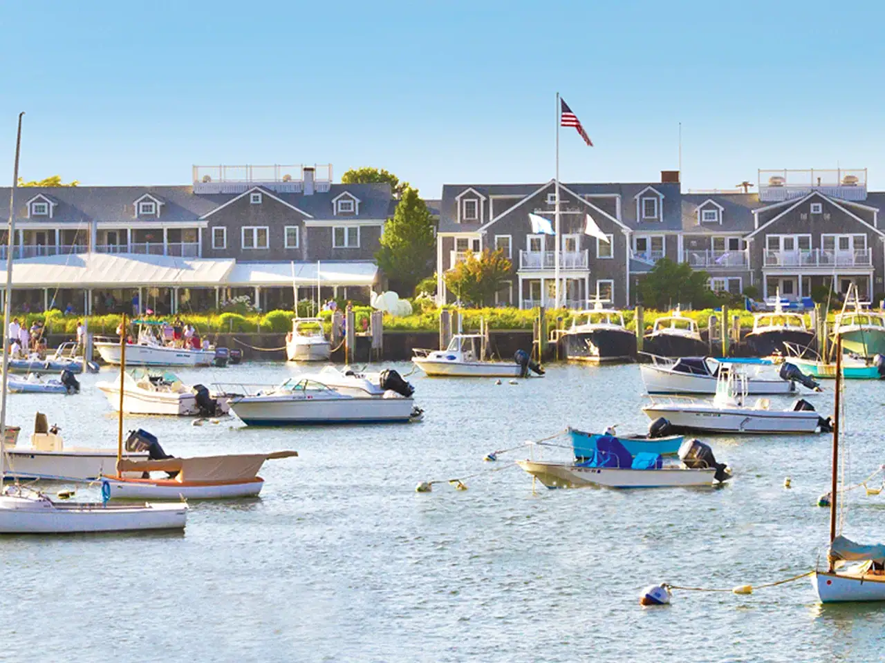 Boats docked in a marina with waterfront buildings and an American flag in the background.